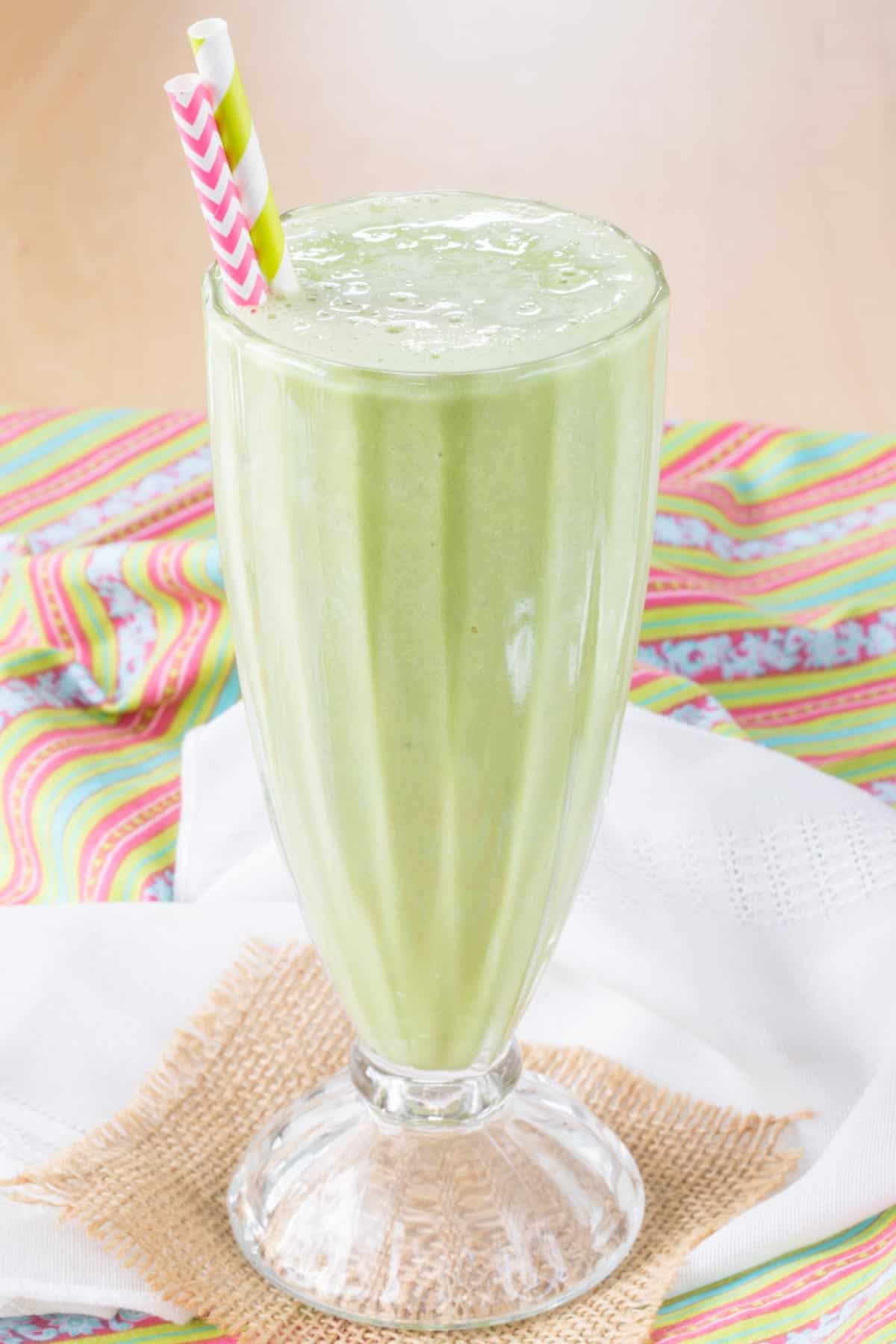A green mint smoothie in a milkshake glass with two striped straws on top of cloth napkins and a small piece of burlap.