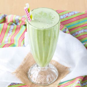 A green mint smoothie in a milkshake glass with two striped straws on top of a colorful striped cloth napkin, a white napkin, and a small piece of burlap.