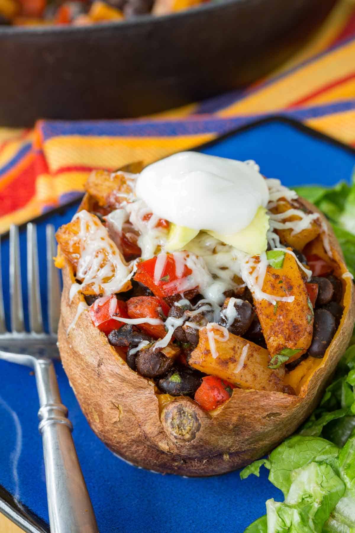 A Mexican sweet potato stuffed with a black bean mixture and taco toppings on a blue plate with a fork.