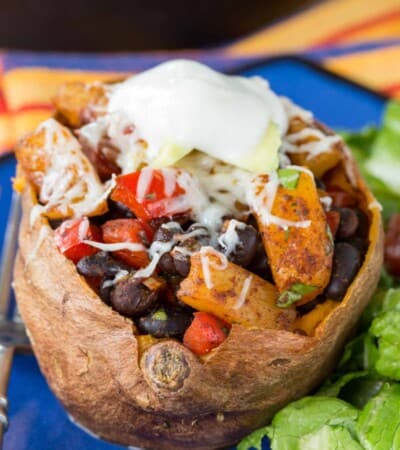 A Mexican Pineapple Black Bean Stuffed Baked Sweet Potato on a blue plate topped with cheese, avocado, and sour cream.