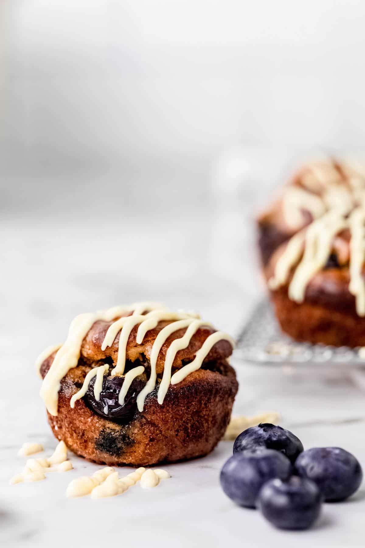 Close up of a flourless blueberry mini muffin drizzled with maple cream cheese next to blueberries, with more muffins on a wire rack in the background.