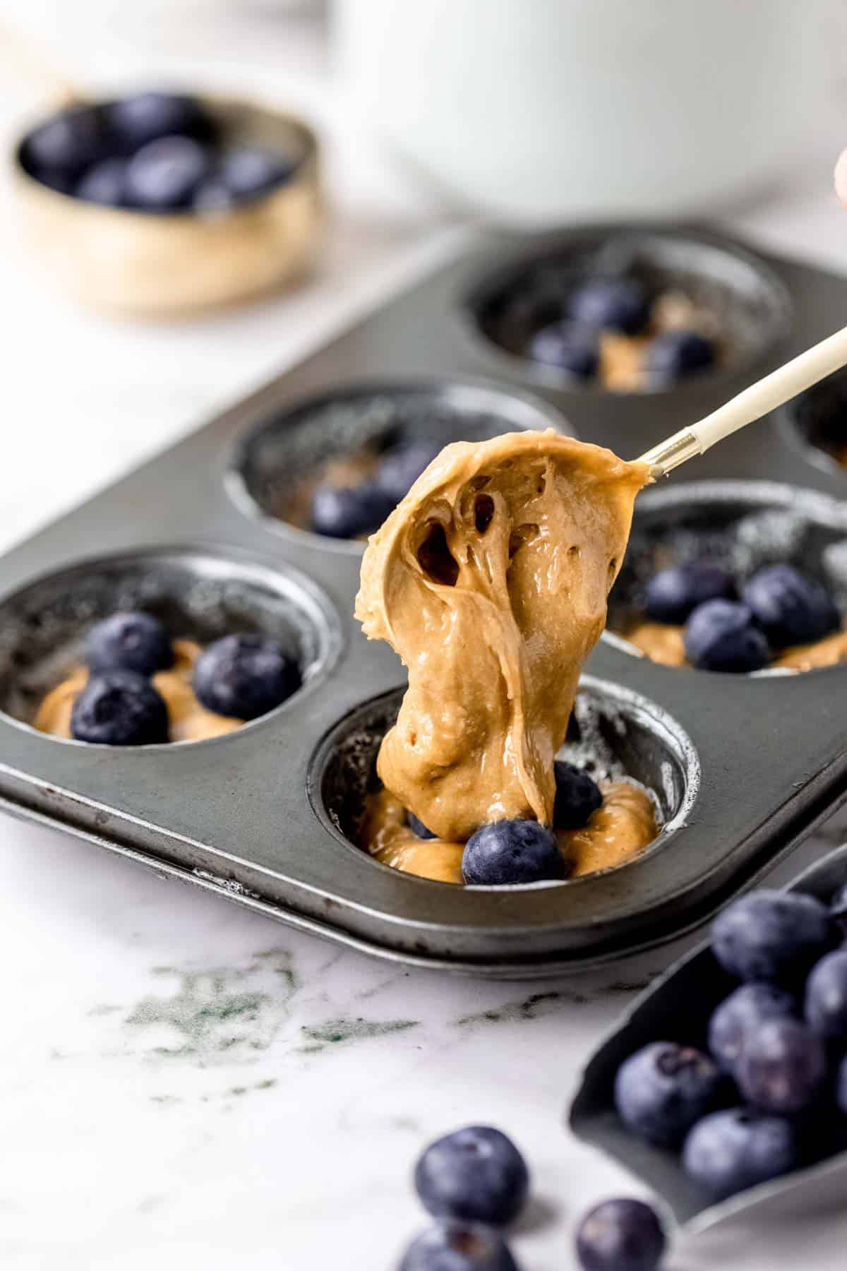 A spoon drops flourless muffin batter over top of another layer of batter and blueberries inside the well of a muffin pan.