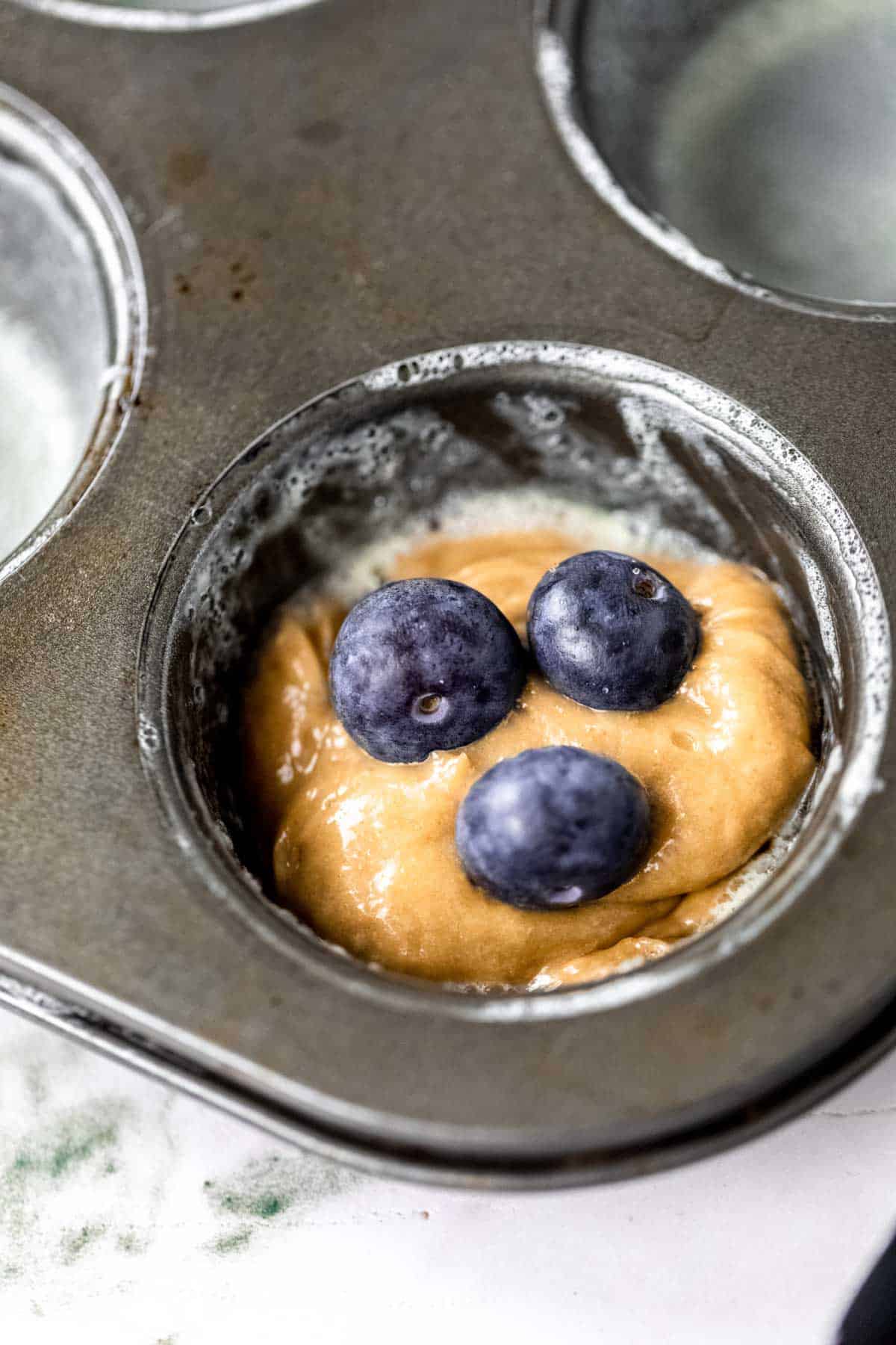 Close up of blueberries placed on top of flourless muffin batter inside the well of a muffin pan.