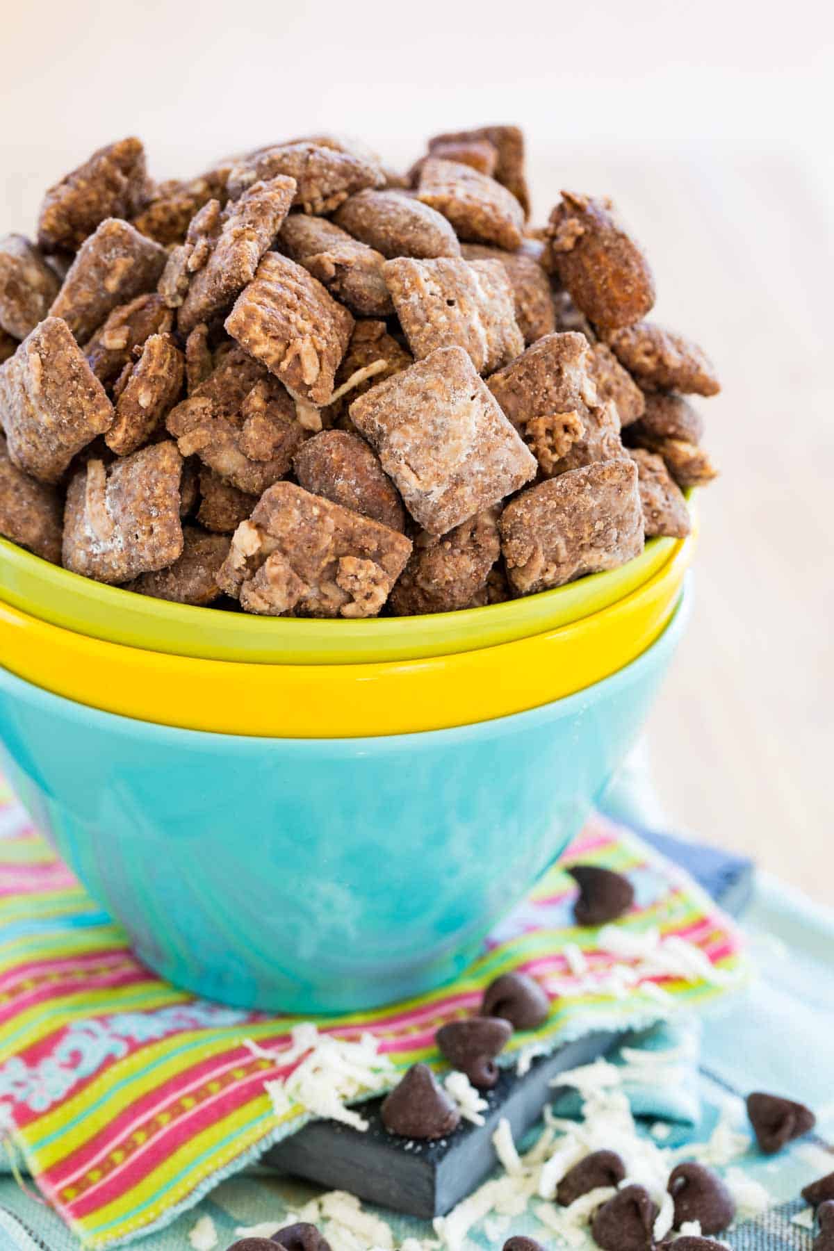 Three blue, yellow, and green bowls stacked together with puppy chow snack mix in the top one and coconut and chocolate chips scattered on the table.