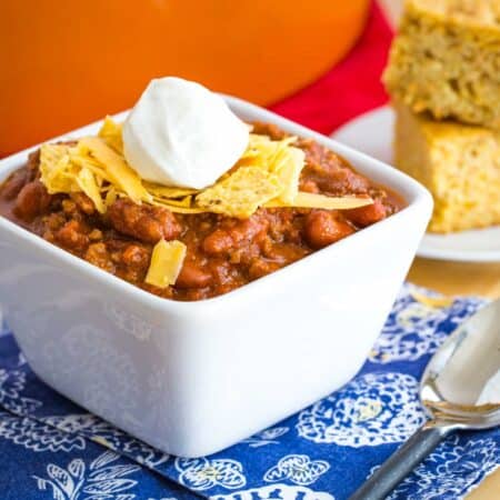chili in a bowl with crushed tortilla chips, shredded cheese, and sour cream on top and a plate of cornbread next to it