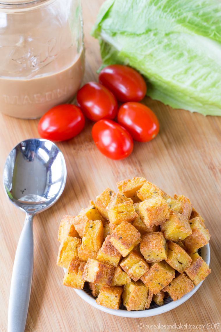 A small bowl of baked polenta croutons with lettuce, tomatoes, and a jar of dressing in the background