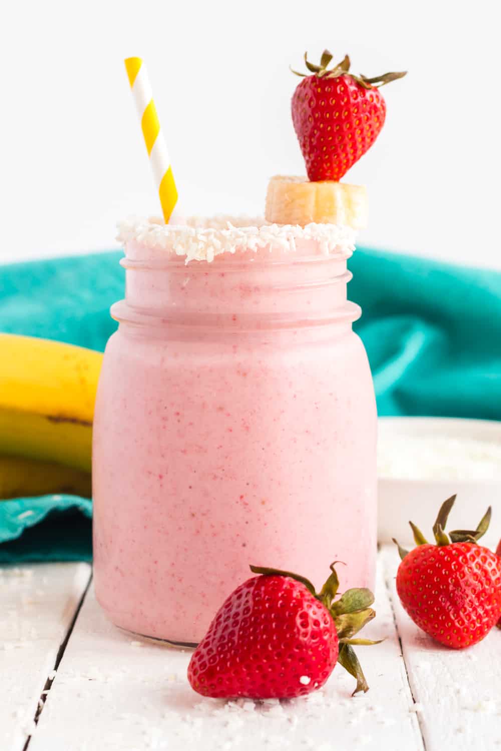 A strawberry banana smoothie in a glass jar with a striped straw, and garnished with a strawberry, banana slice, and shredded coconut on the rim.