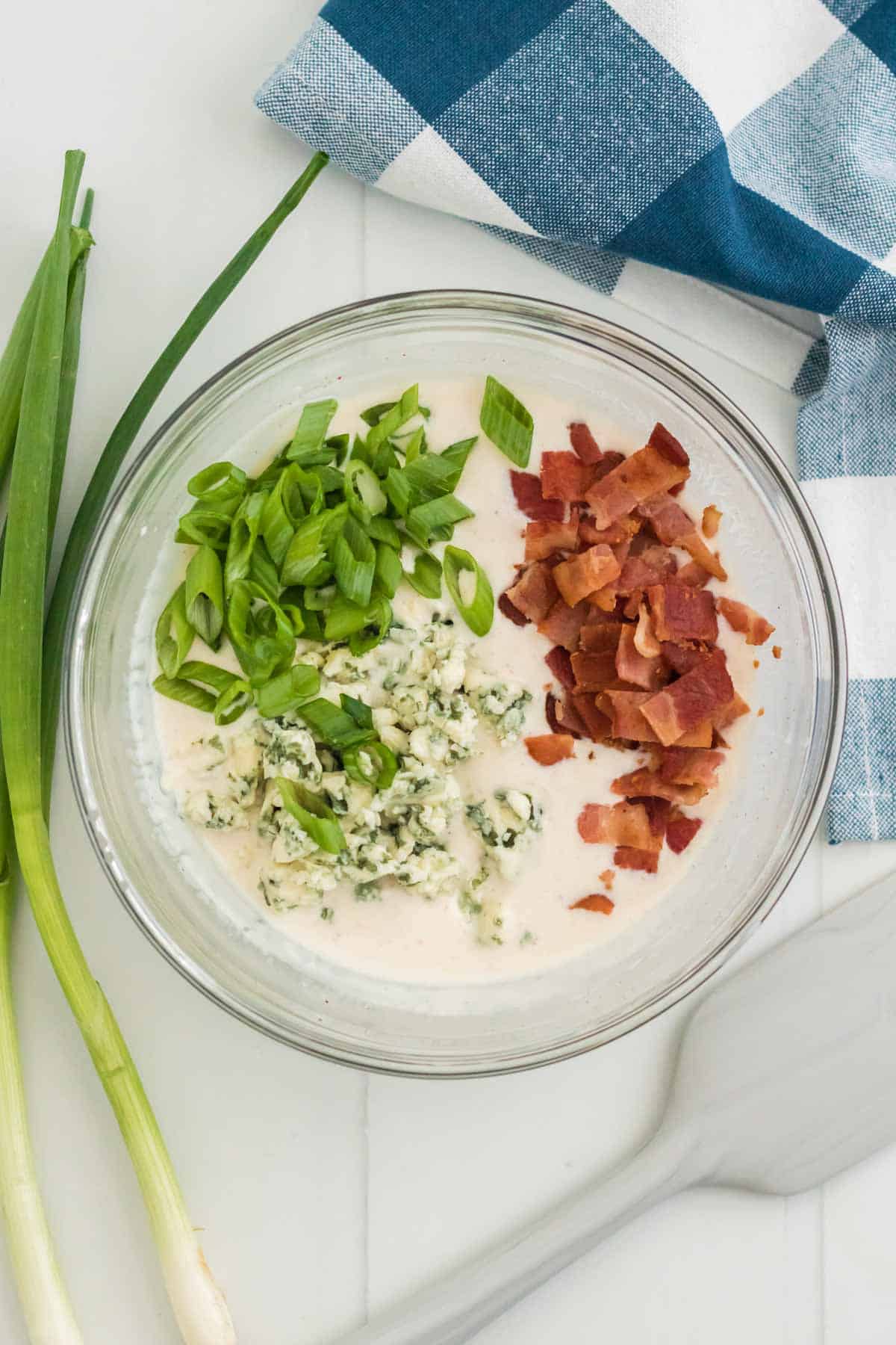 Chopped bacon, blue cheese, and scallions are added into a bowl with potato salad dressing.