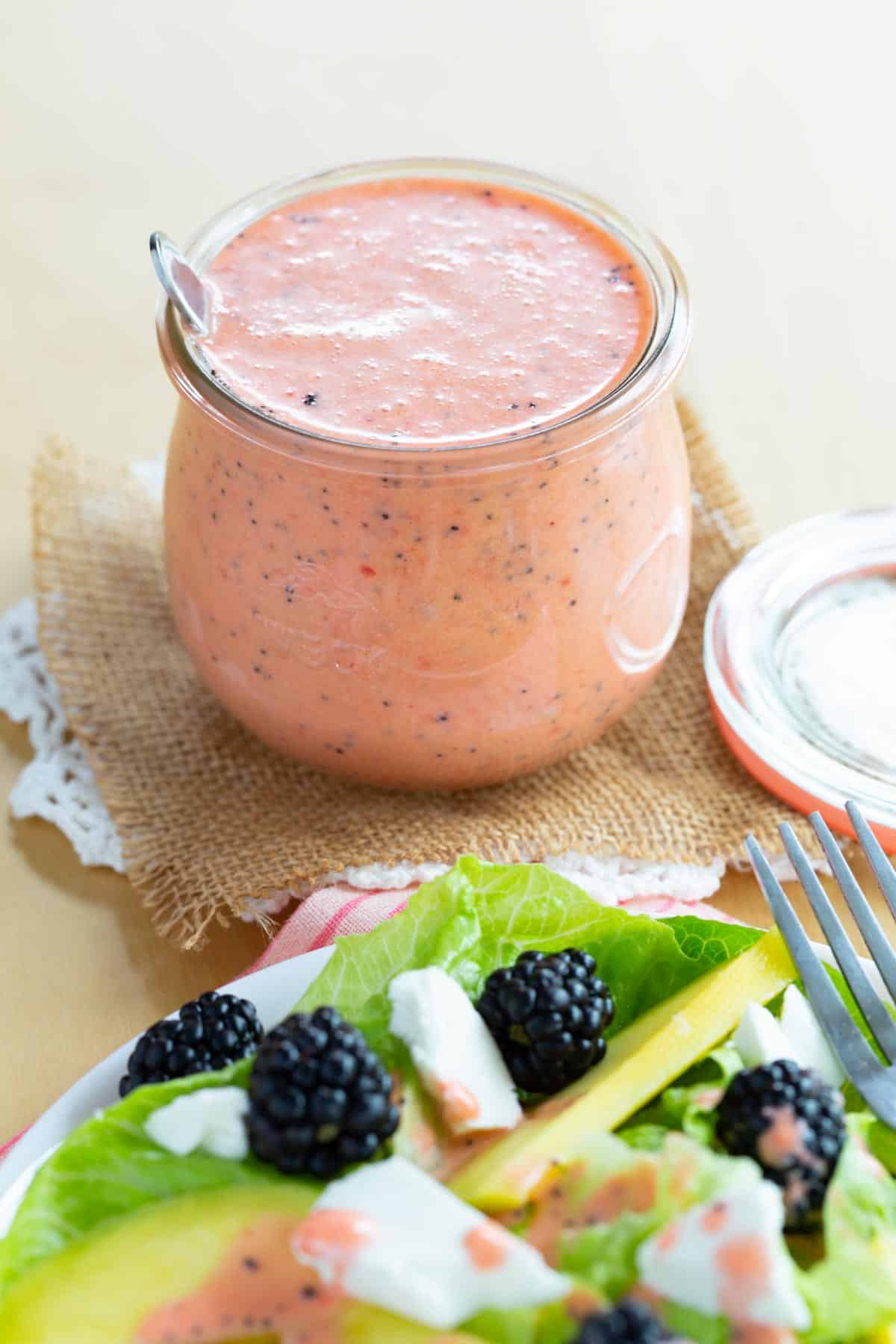 A spoon in a jar of strawberry poppy seed dressing next to a plate of salad.