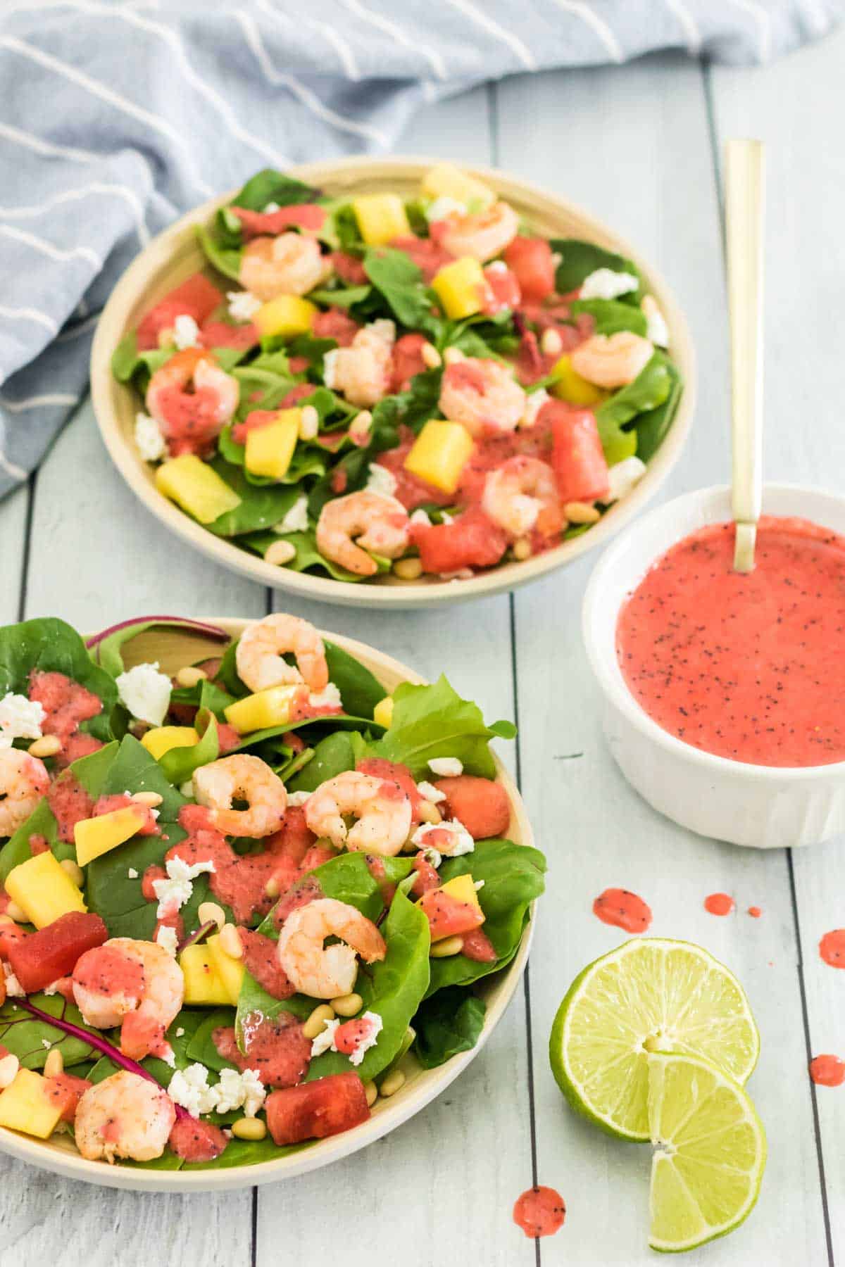 two bowls of grilled shrimp salad with a bowl of strawberry poppy seed dressing and sliced limes next to them