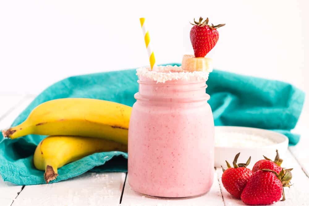 Small glass jar with Coconut Strawberry Banana Smoothie and a turquoise napkin with fruit in the background