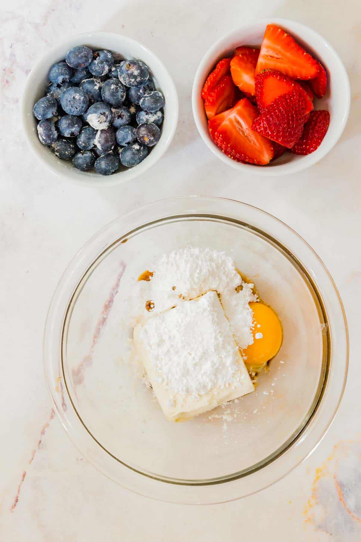 The ingredients for the cream cheese layer combined in a bowl, next to bowls of blueberries and strawberries.