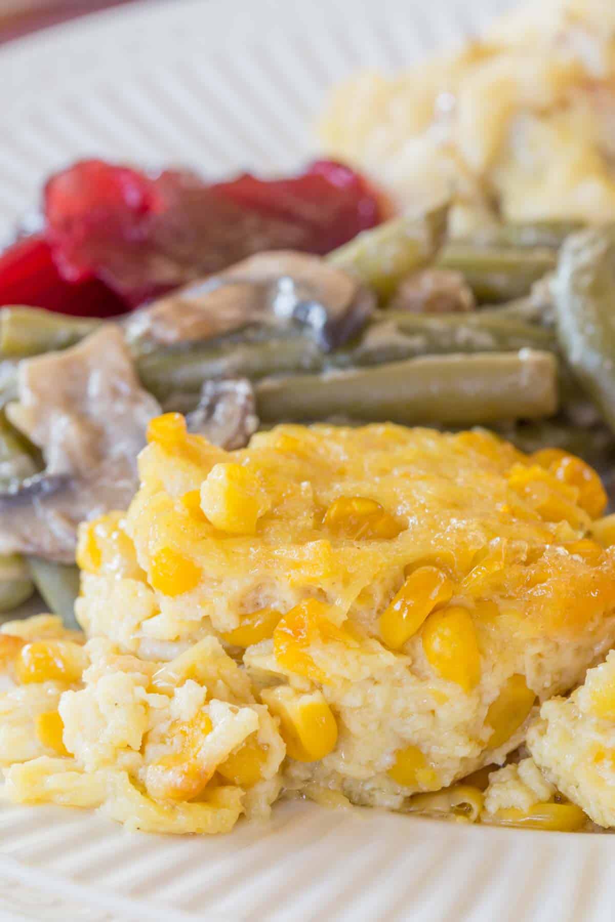 A scoop of corn pudding on a plate with green beans, cranberry sauce, and mashed potatoes.