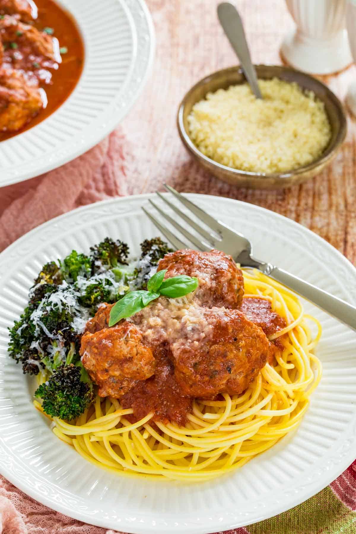 A plateful of spaghetti and Italian meatballs with a side of roasted broccoli, topped with parmesan next to a fork.