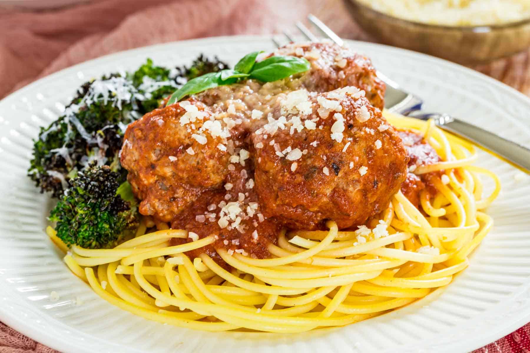 A plateful of spaghetti and gluten-free Italian meatballs with a side of roasted broccoli, topped with parmesan next to a fork.