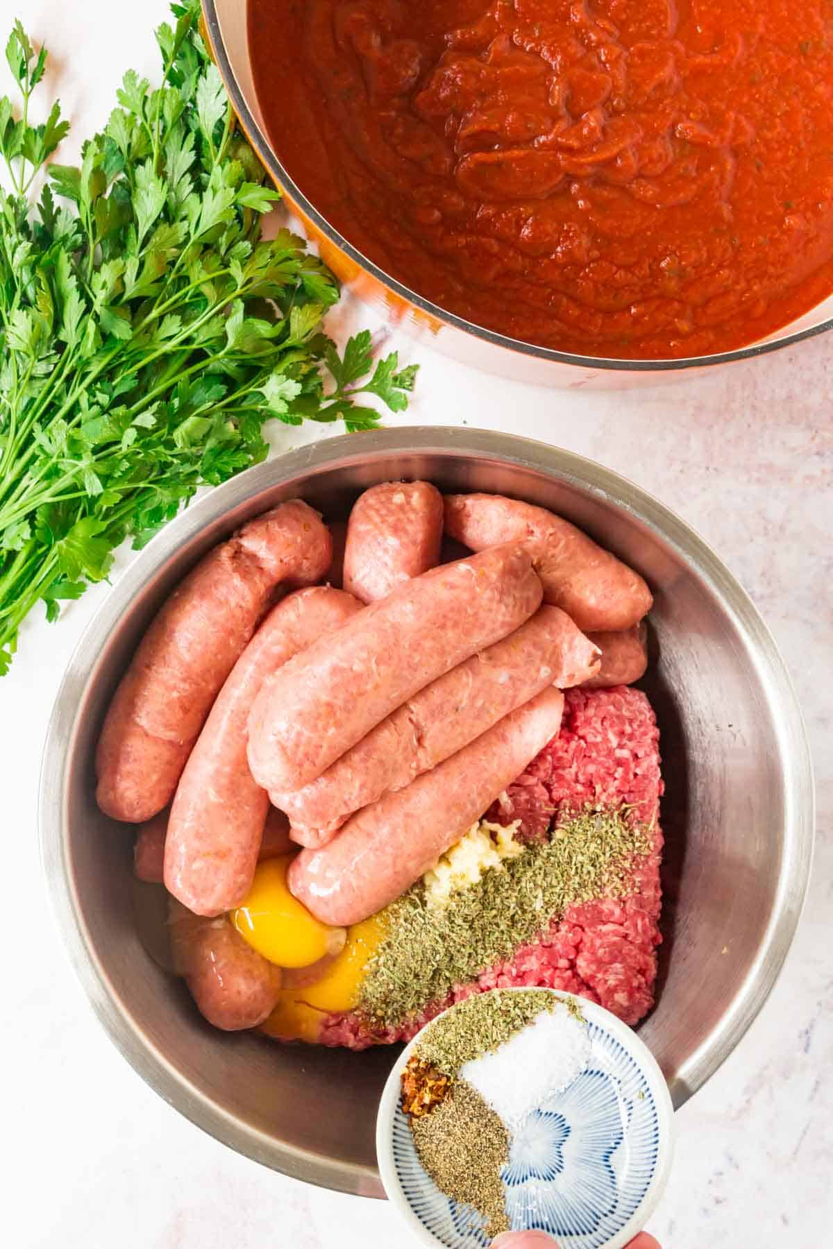 Seasonings are added to a metal bowl with the ingredients for Italian meatballs.