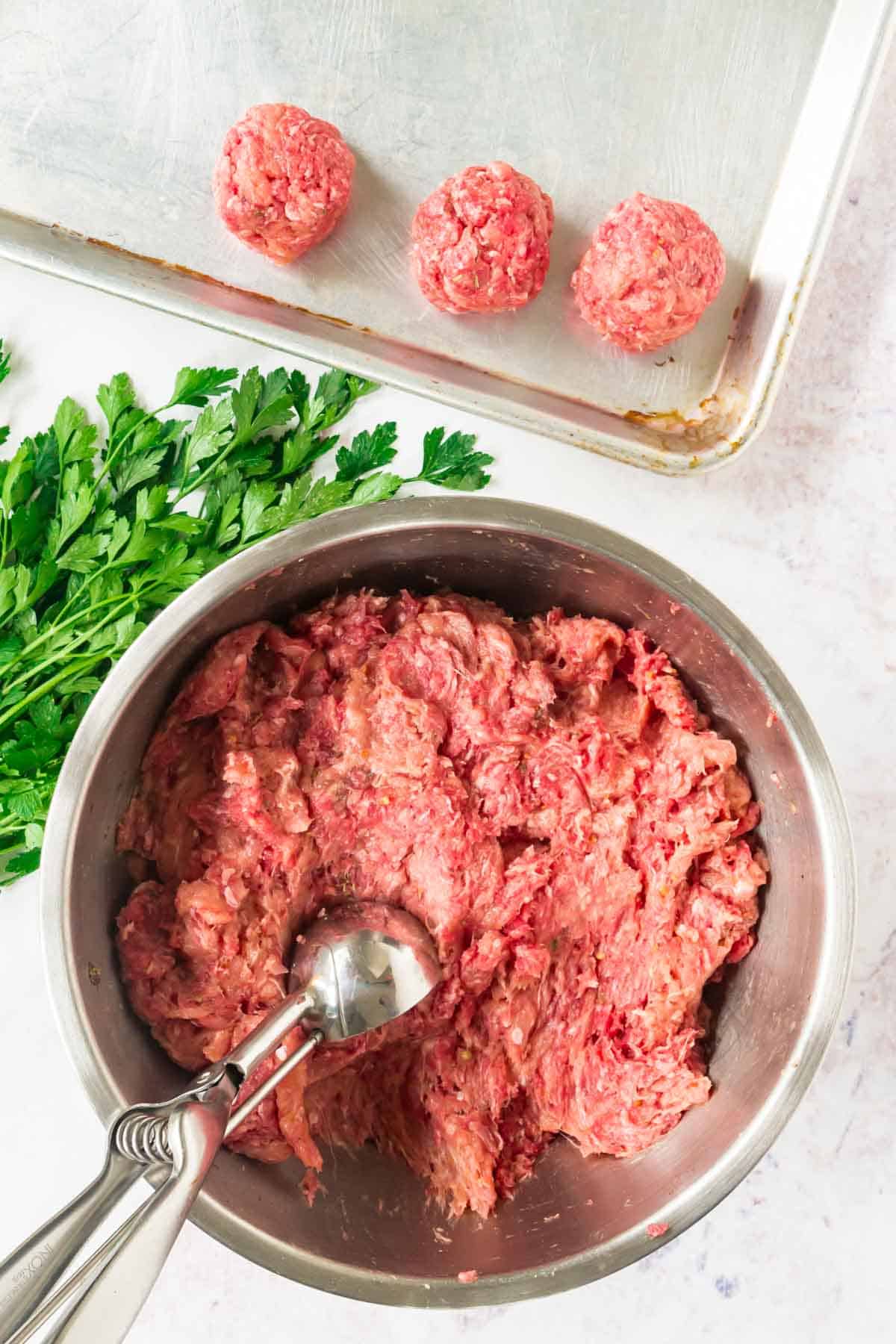 A scoop is used to portion the meat mixture into meatballs on a baking sheet.
