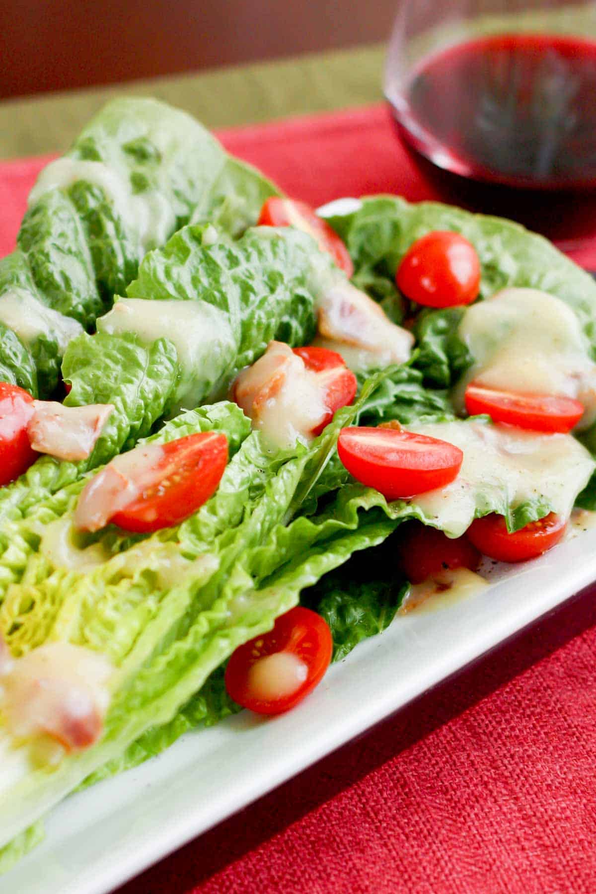 Half a head of romaine lettuce drizzled with hot maple bacon dressing and topped with halved grape tomatoes next to a glass of red wine.