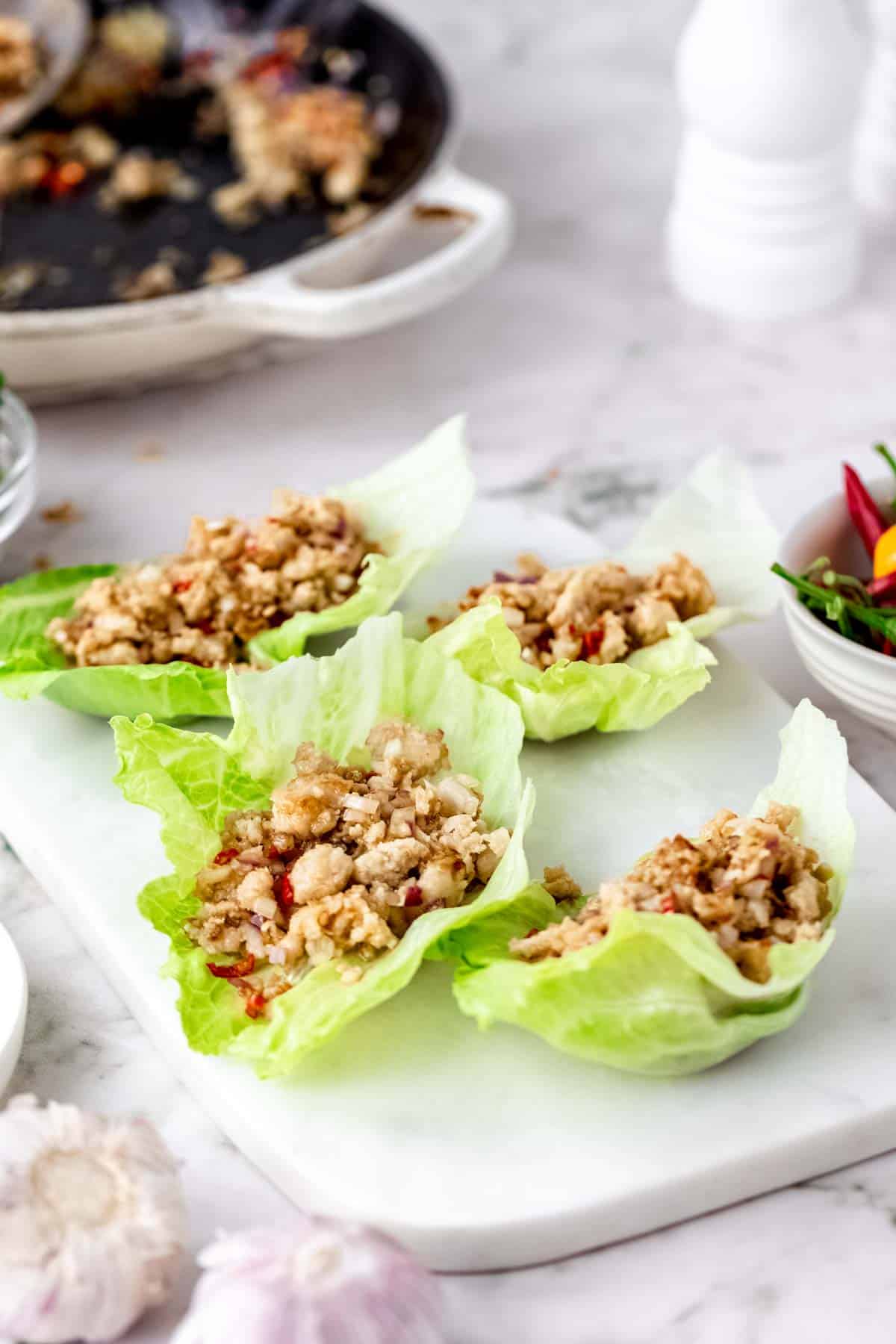 Thai larb chicken lettuce wraps on a white stone cutting board, next to a bowl of chili peppers with a skillet in the background.