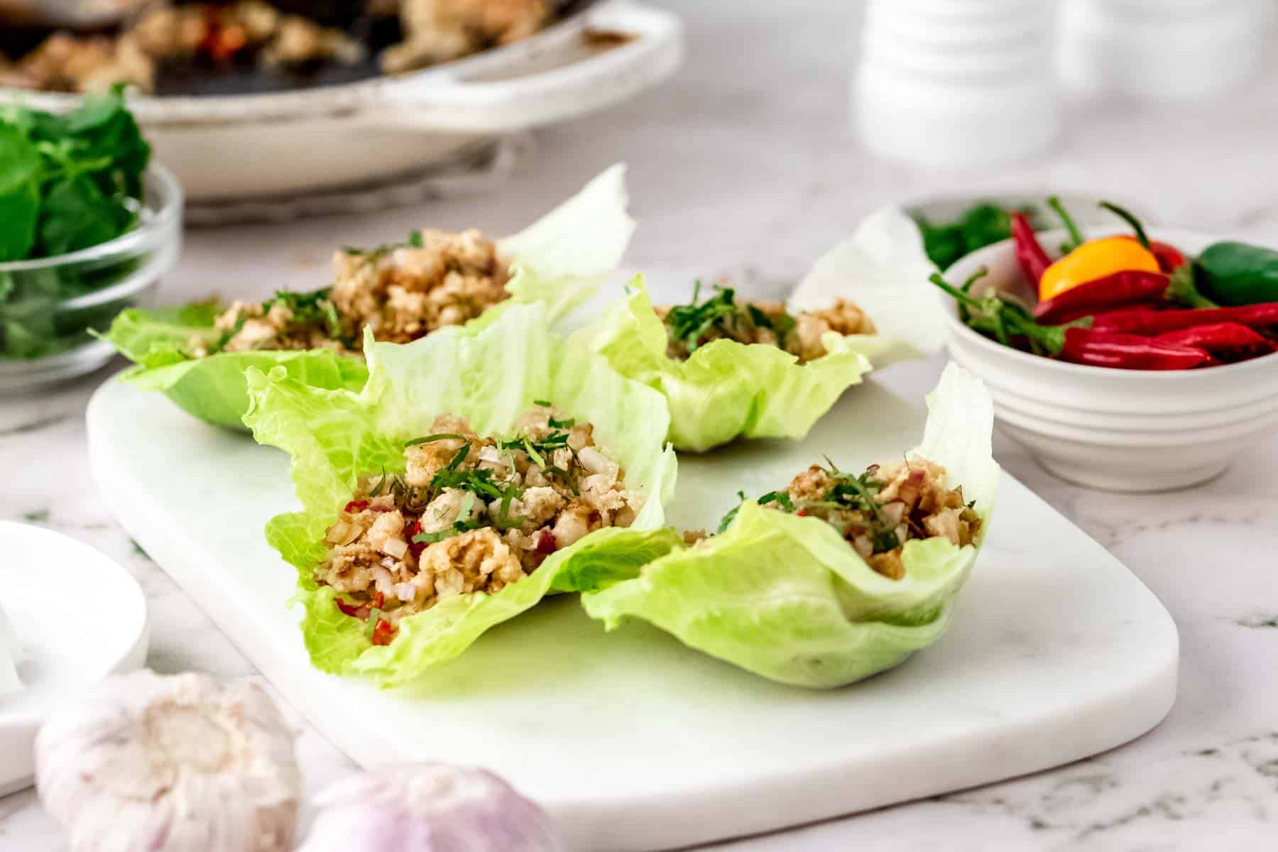Thai larb chicken lettuce wraps on a white stone cutting board, next to a bowl of chili peppers and garlic bulbs.