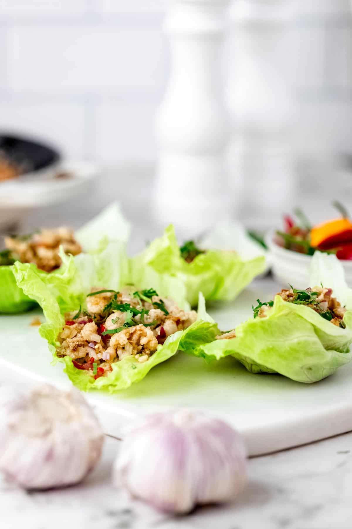 Thai larb chicken lettuce wraps on a white stone cutting board, next to a bowl of chili peppers and garlic bulbs.