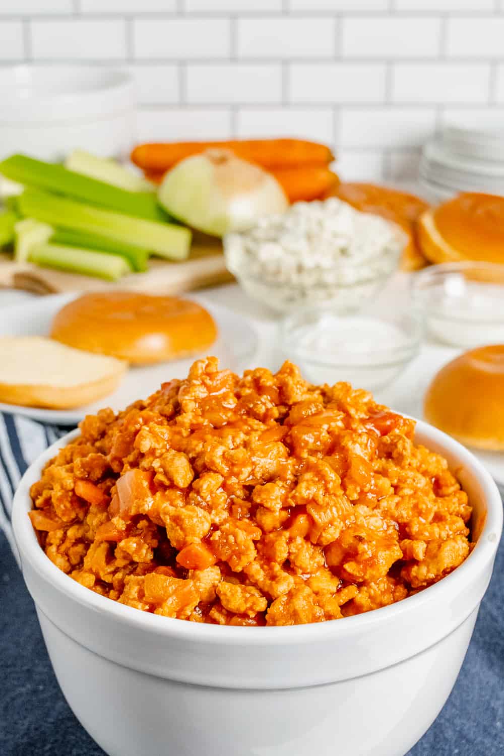 Buffalo chicken sloppy joes ground chicken mixture in a bowl with rolls and toppings behind it
