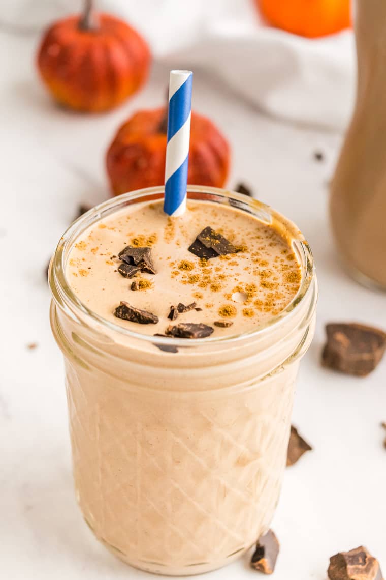 A Chocolate Pumpkin Smoothie in a jelly jar with a sprinkle of pumpkin pie spice and bits of chocolate on top and a blue and white striped straw