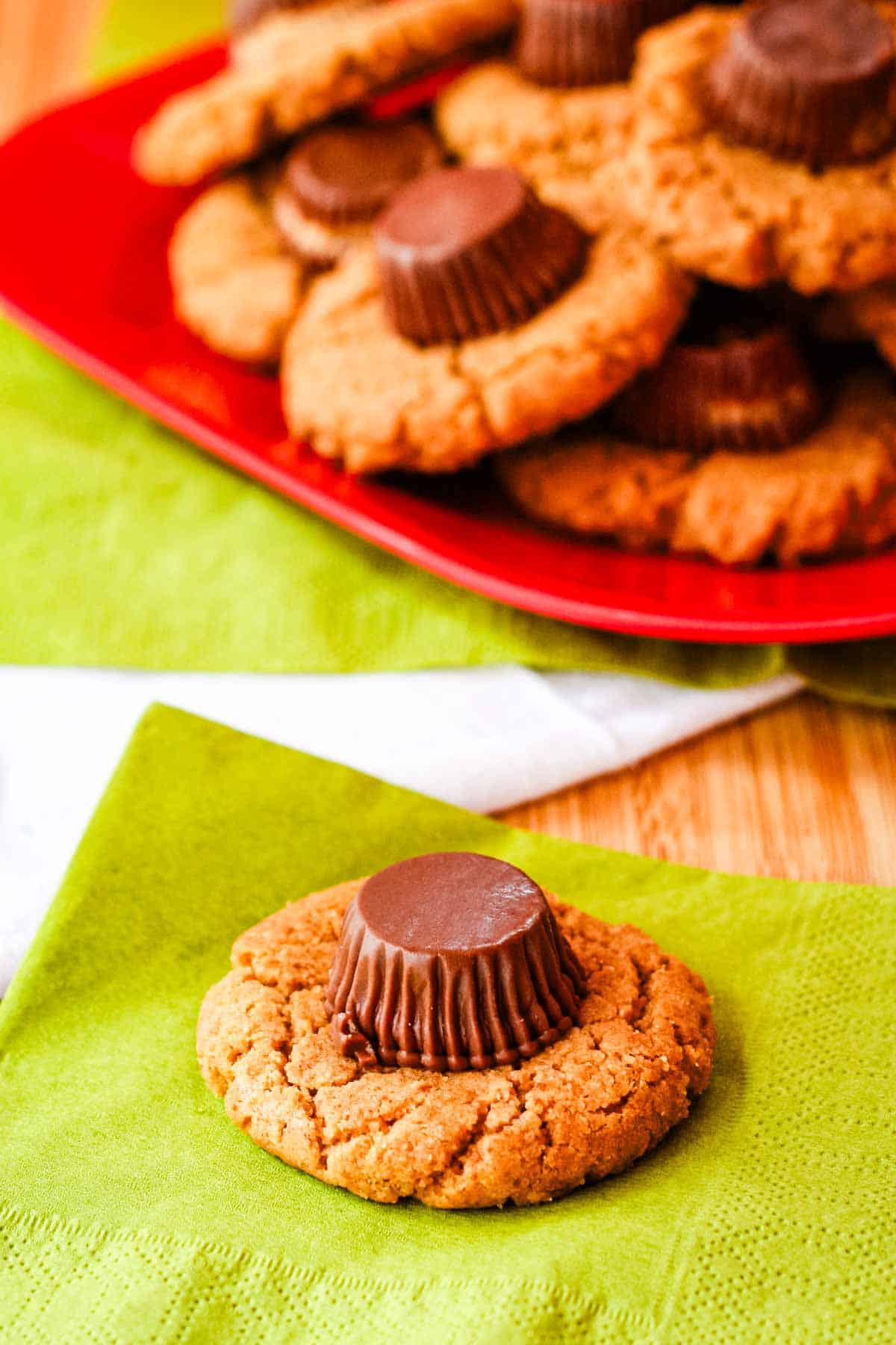 A peanut butter cookie topped with a Reese's peanut butter cup on a green napkin with more cookies on a red plate in the background.