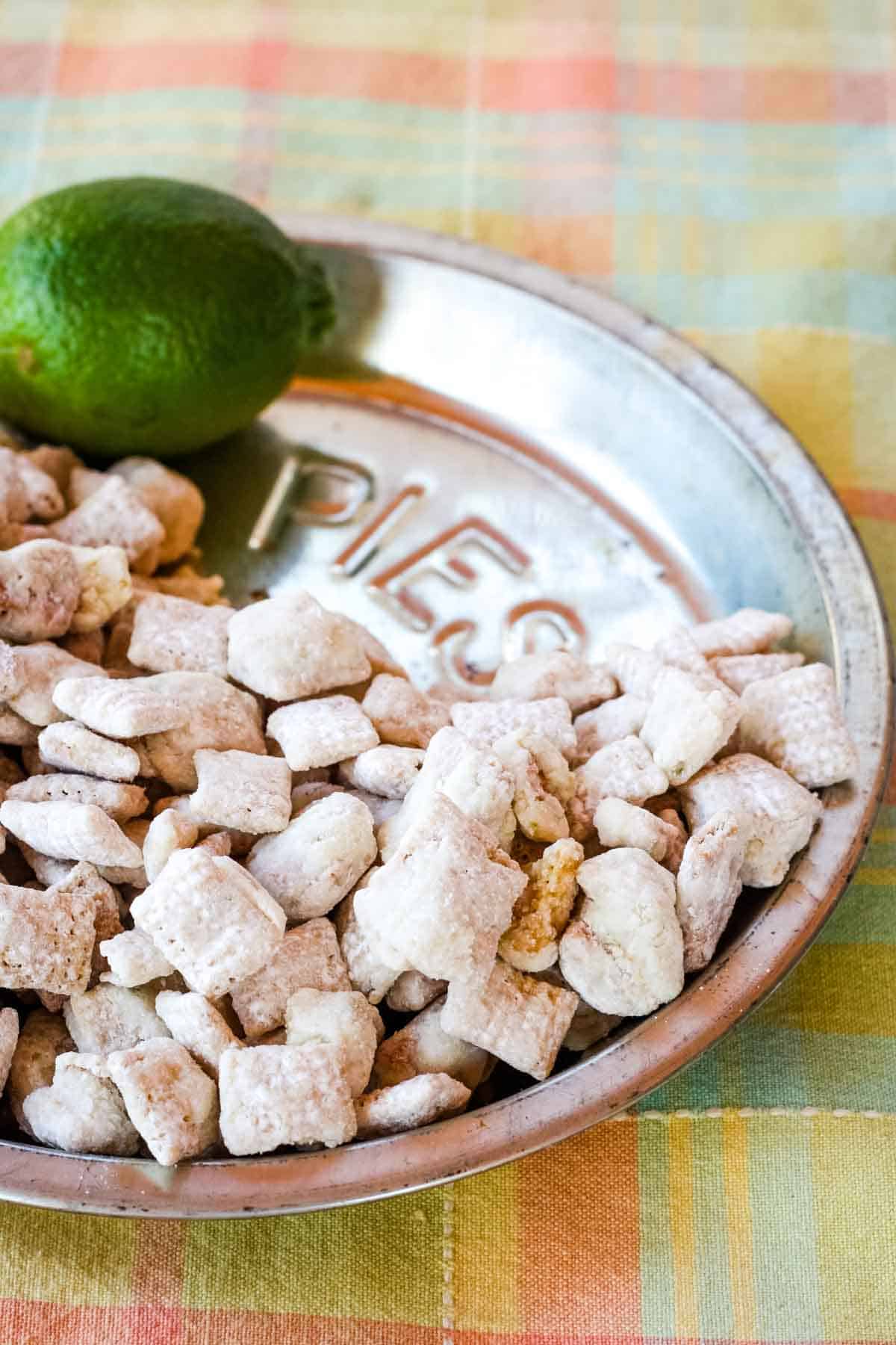 White chocolate puppy chow snack mix in a metal pie plate with a lime on top of a pastel plaid placemat.