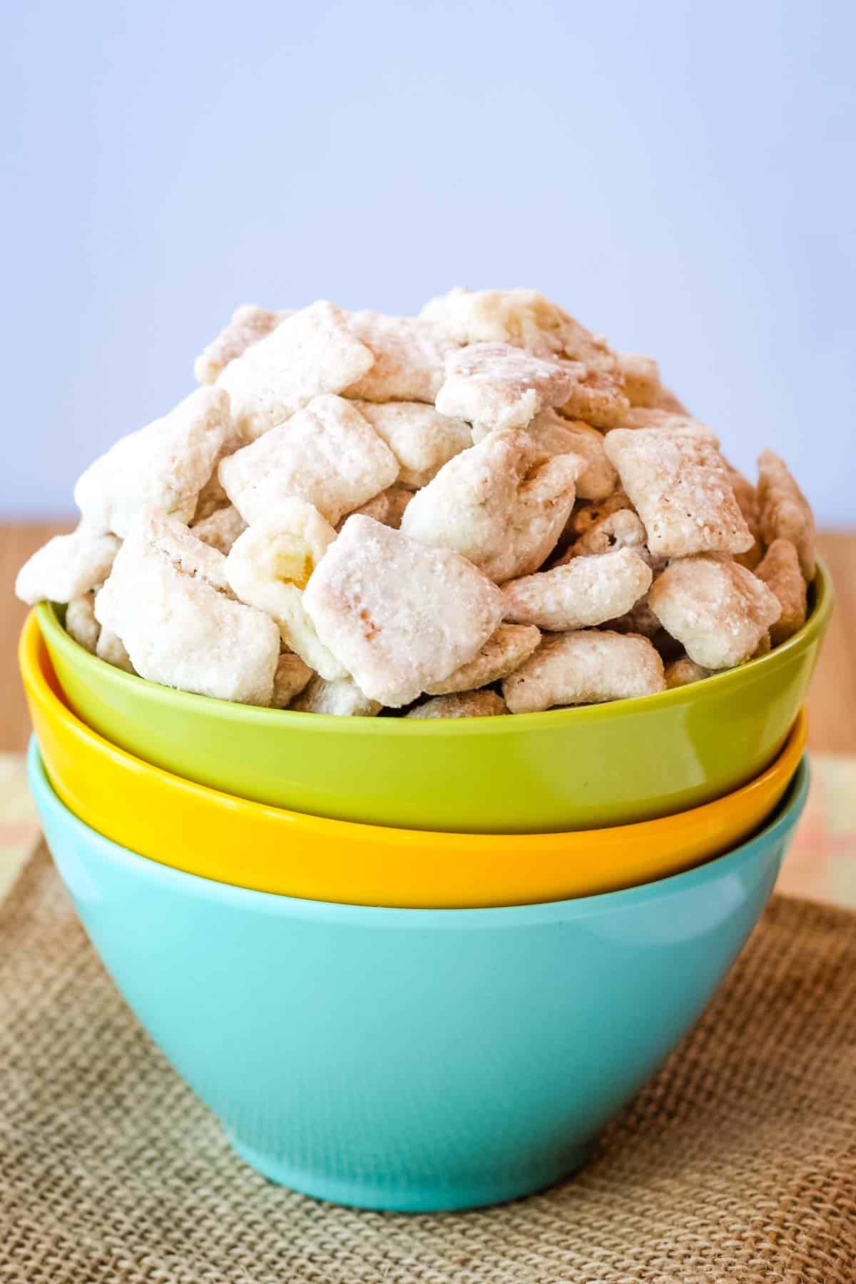 Three bowls stacked up with the top one filled with Key Lime Pie White Chocolate Puppy Chow on top of a piece of burlap with a blue background.