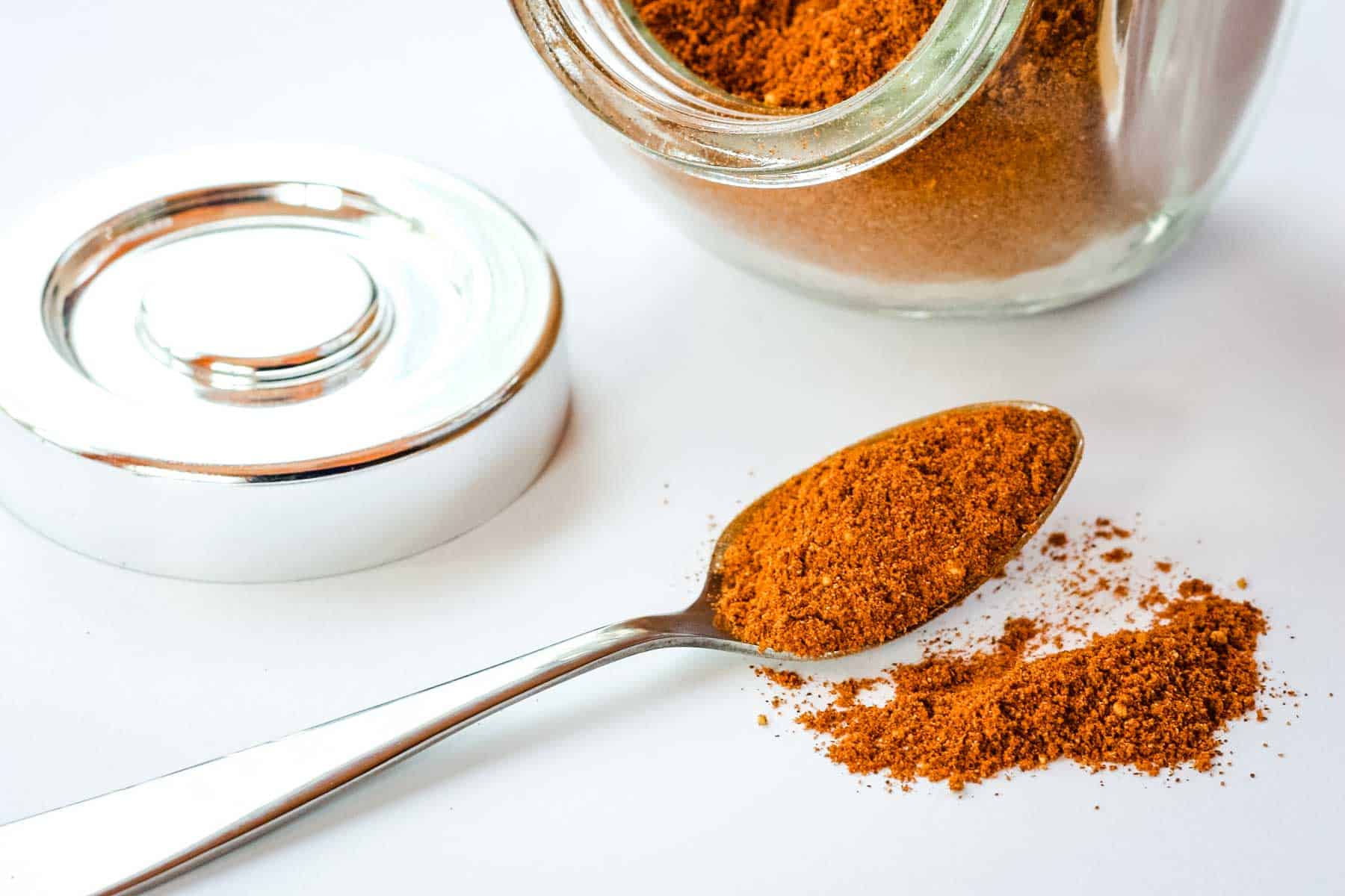 A spoon filled with a spice mix with some falling off onto a white table.