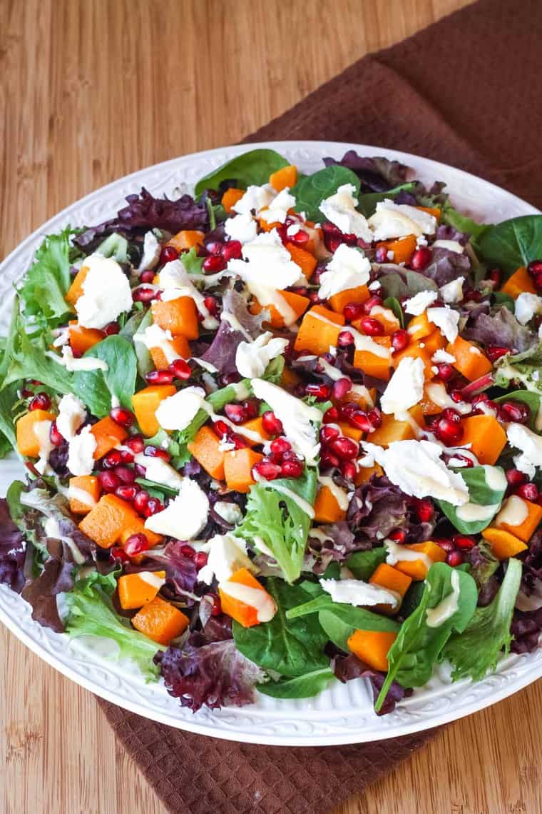 Large dinner plate filled with baby greens, roasted butternut squash cubes, pomegranate, and salad dressing