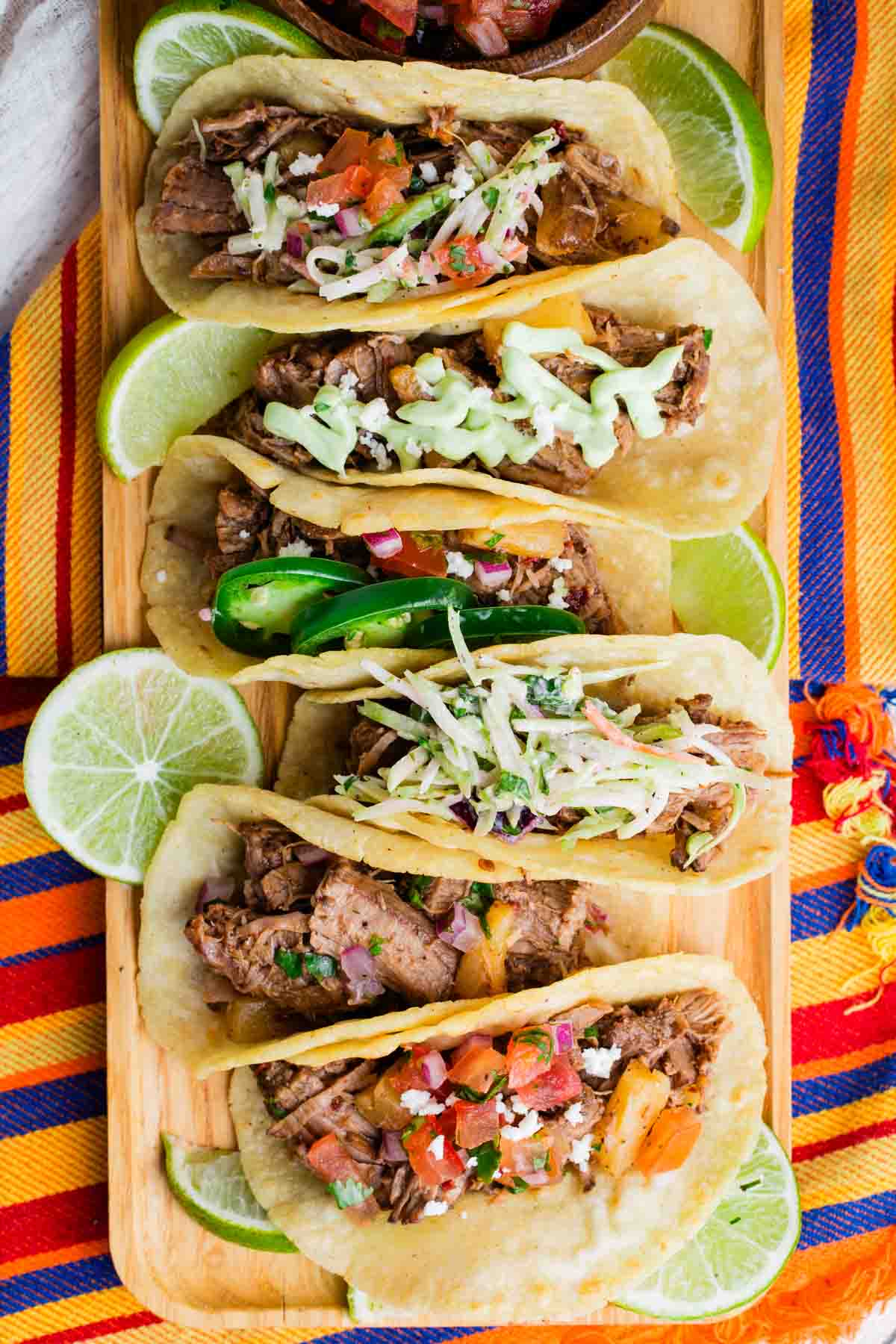 Several beef barbacoa tacos with different toppings on each lined up on a wooden platter.