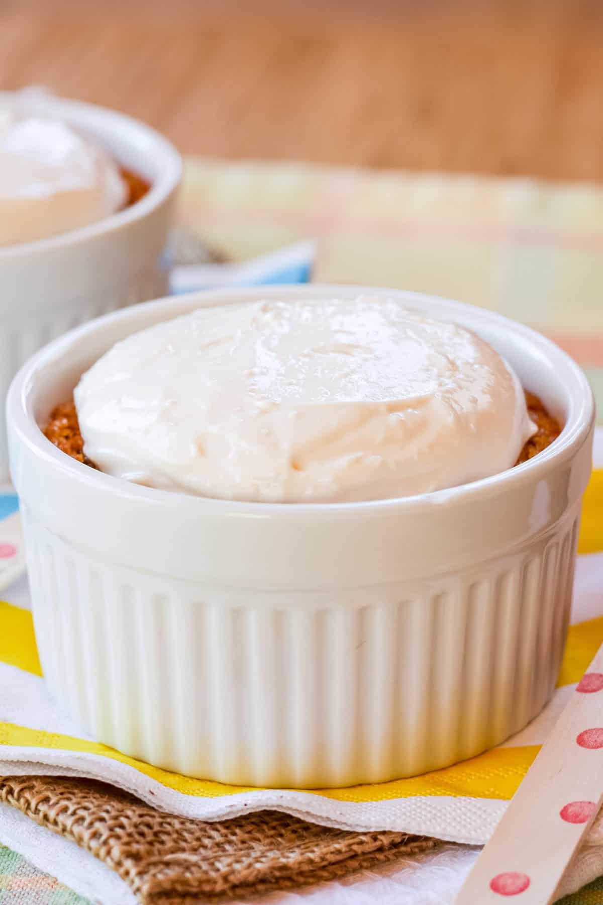 Gluten free carrot cake with cream cheese topping in a white ramekin on top of a stacked white cloth napkin, piece of burlap, and white and yellow striped paper napkin.