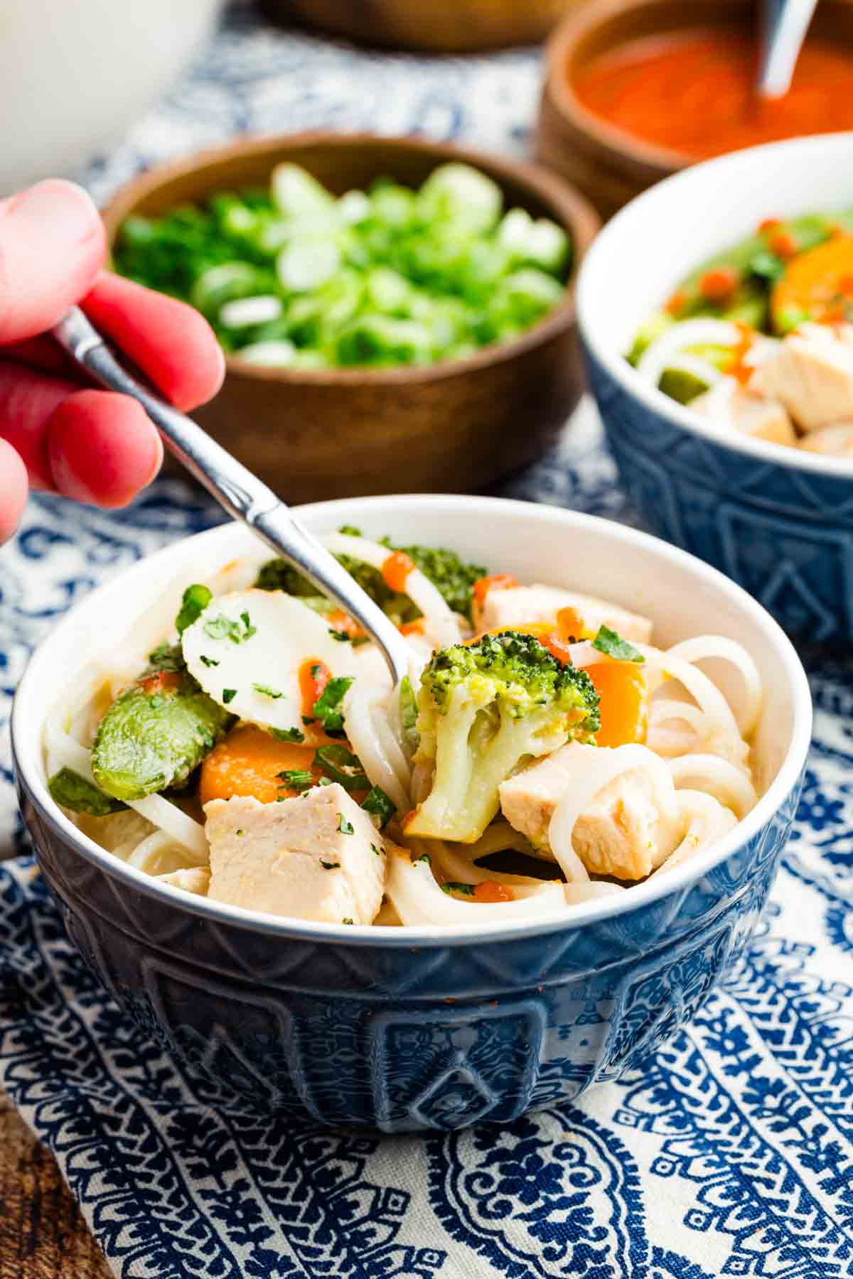 A hand holding a spoon in a blue bowl of Thai chicken noodle soup.