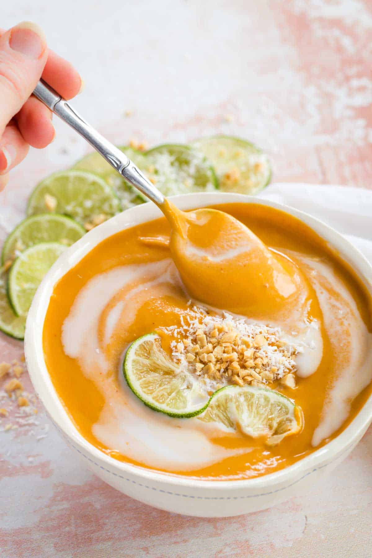 Hand taking a spoonful of Curried Butternut Squash Soup topped with a lime slice plus chopped peanuts and shredded coconut.