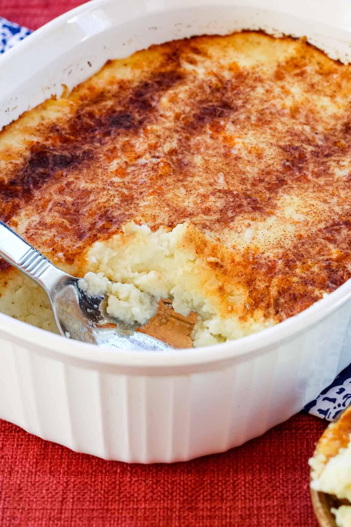 A baking dish of mashed cauliflower casserole with one serving removed and a spoon resting in the dish.