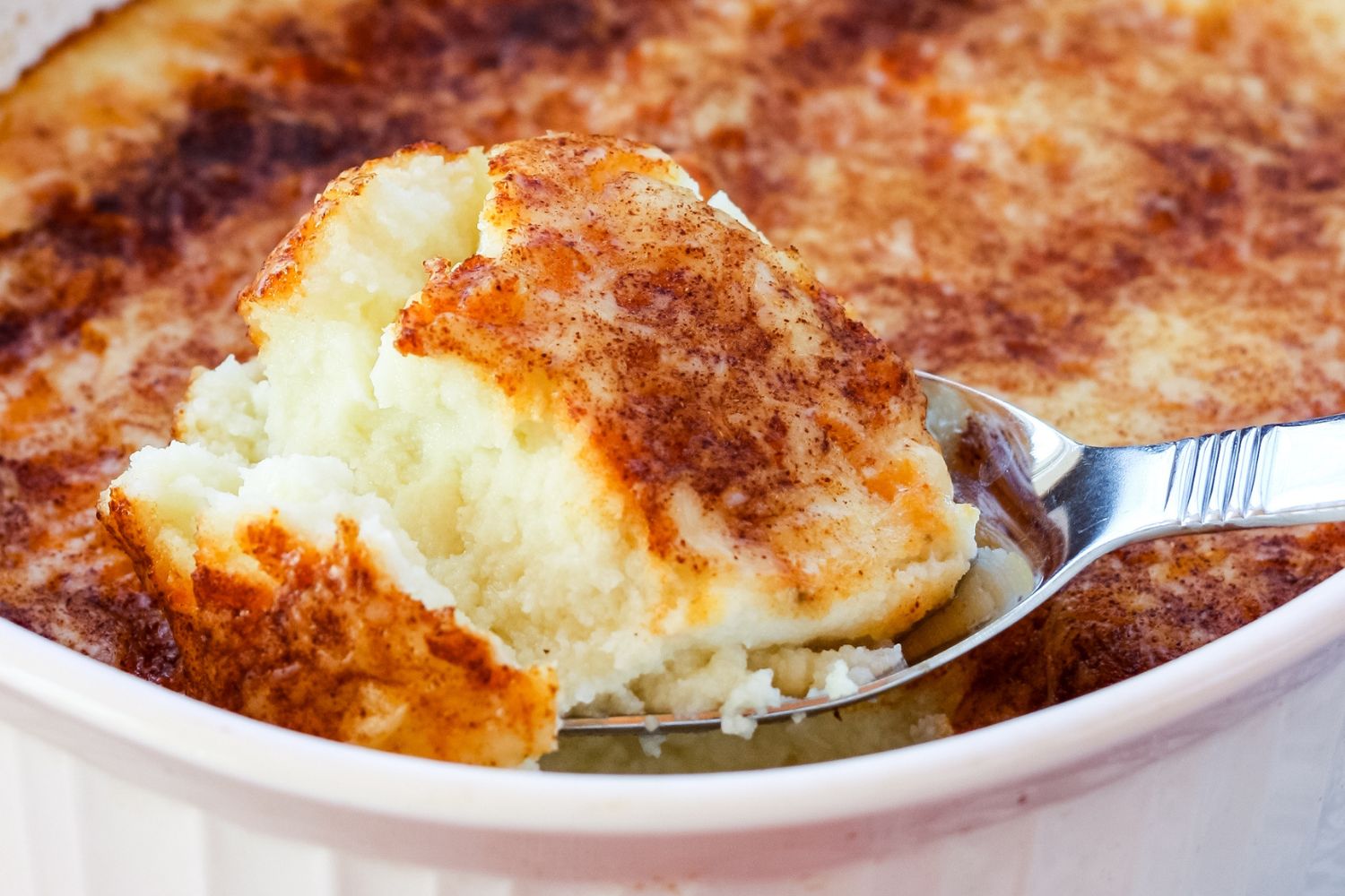 A closeup of a spoon picking up a scoop of cheesy mashed cauliflower from a casserole dish.