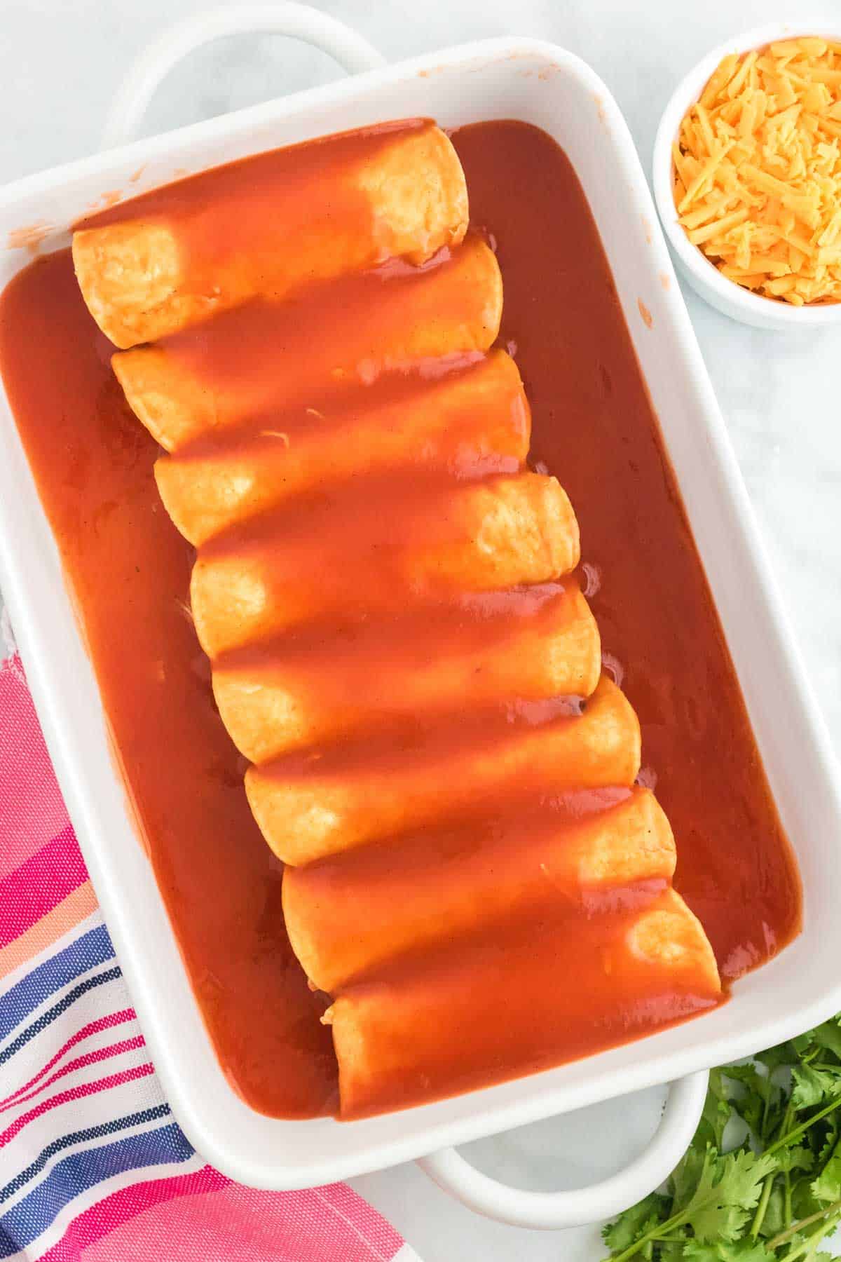 Rolled chicken enchiladas lined up in a baking dish, covered in enchilada sauce.