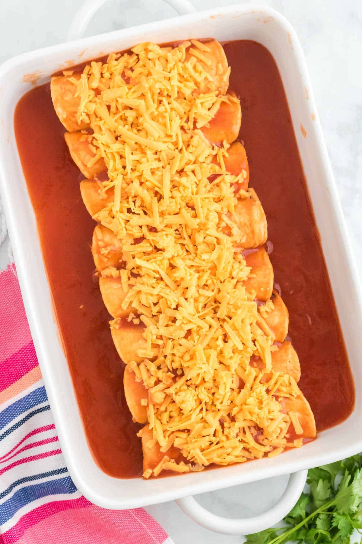 Rolled chicken enchiladas lined up in a baking dish, covered in enchilada sauce and shredded cheese.