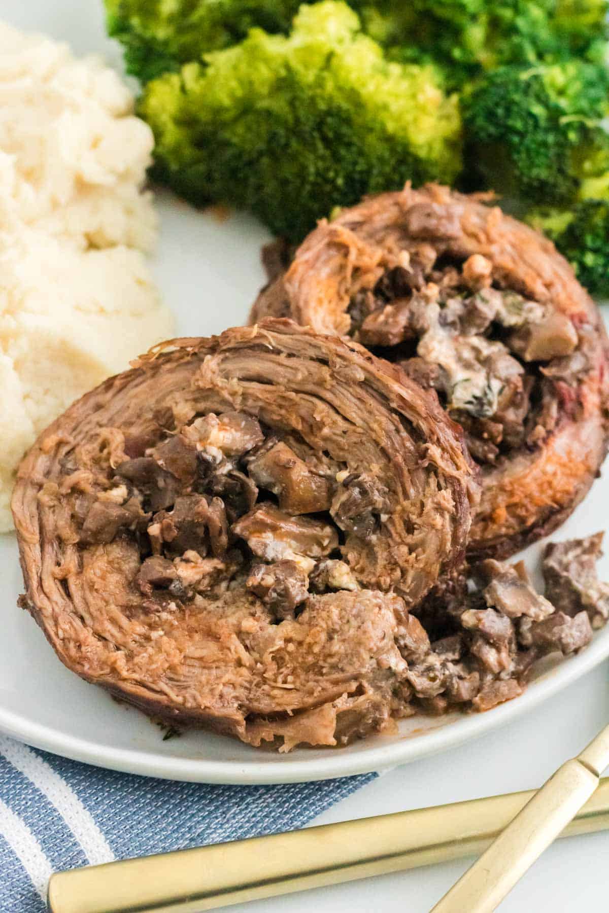 Mushroom stuffed Beef Wellington braciole pinwheels on a plate with broccoli and mashed potatoes.