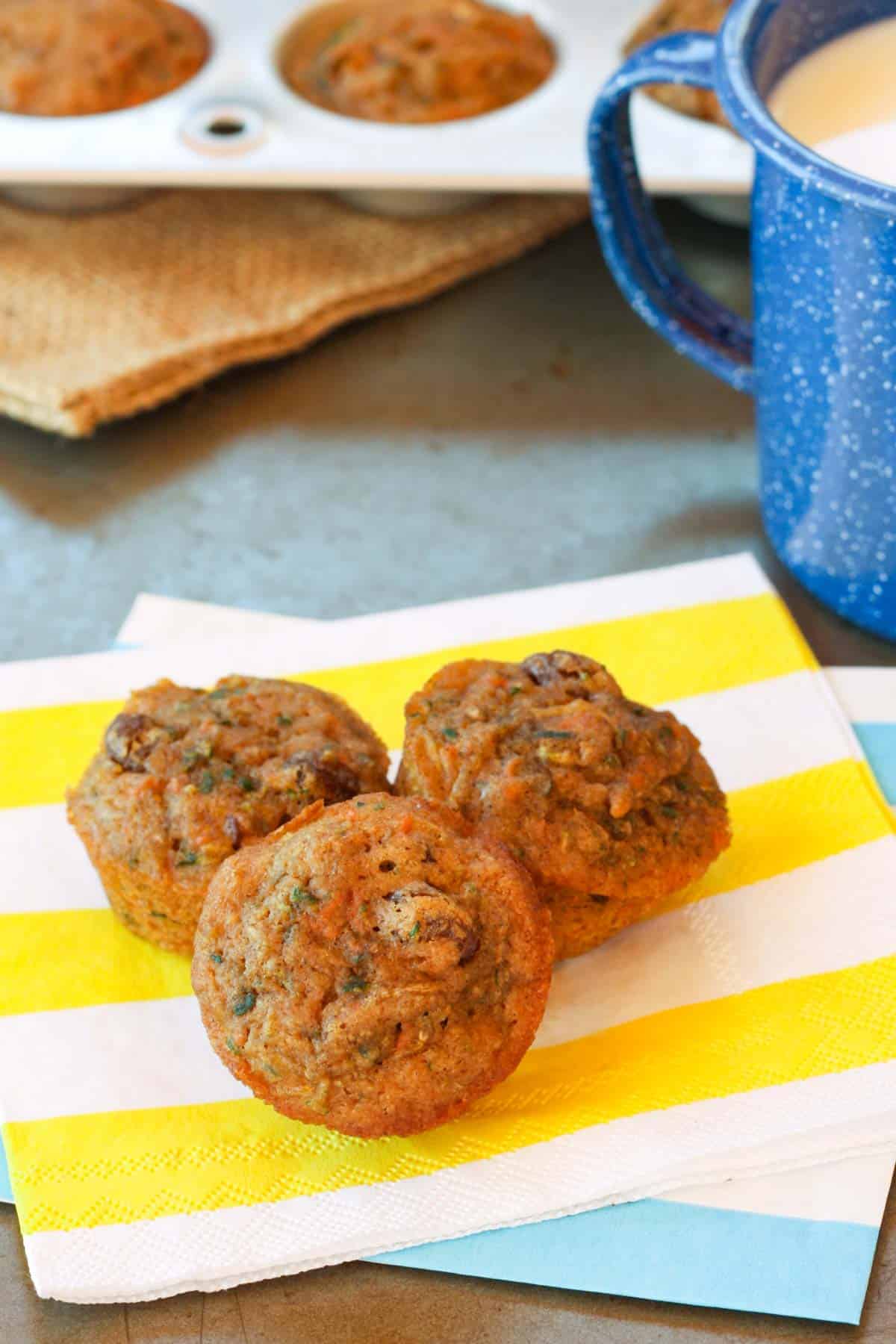 Three carrot zucchini muffins on a yellow and white striped napkin next to a blue cup of milk