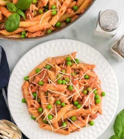 Penne with tomato cream sauce on a plate topped with peas and prosciutto, next to a pan of pasta.