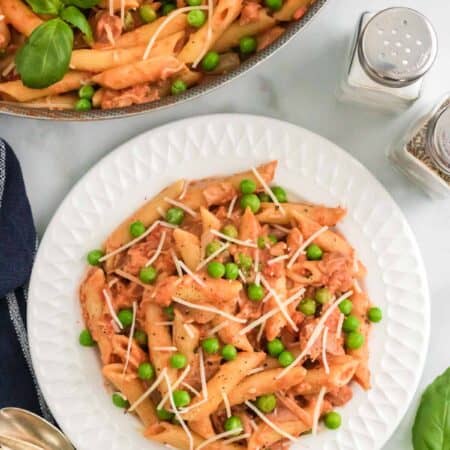 Penne with tomato cream sauce on a plate topped with peas and prosciutto, next to a pan of pasta.