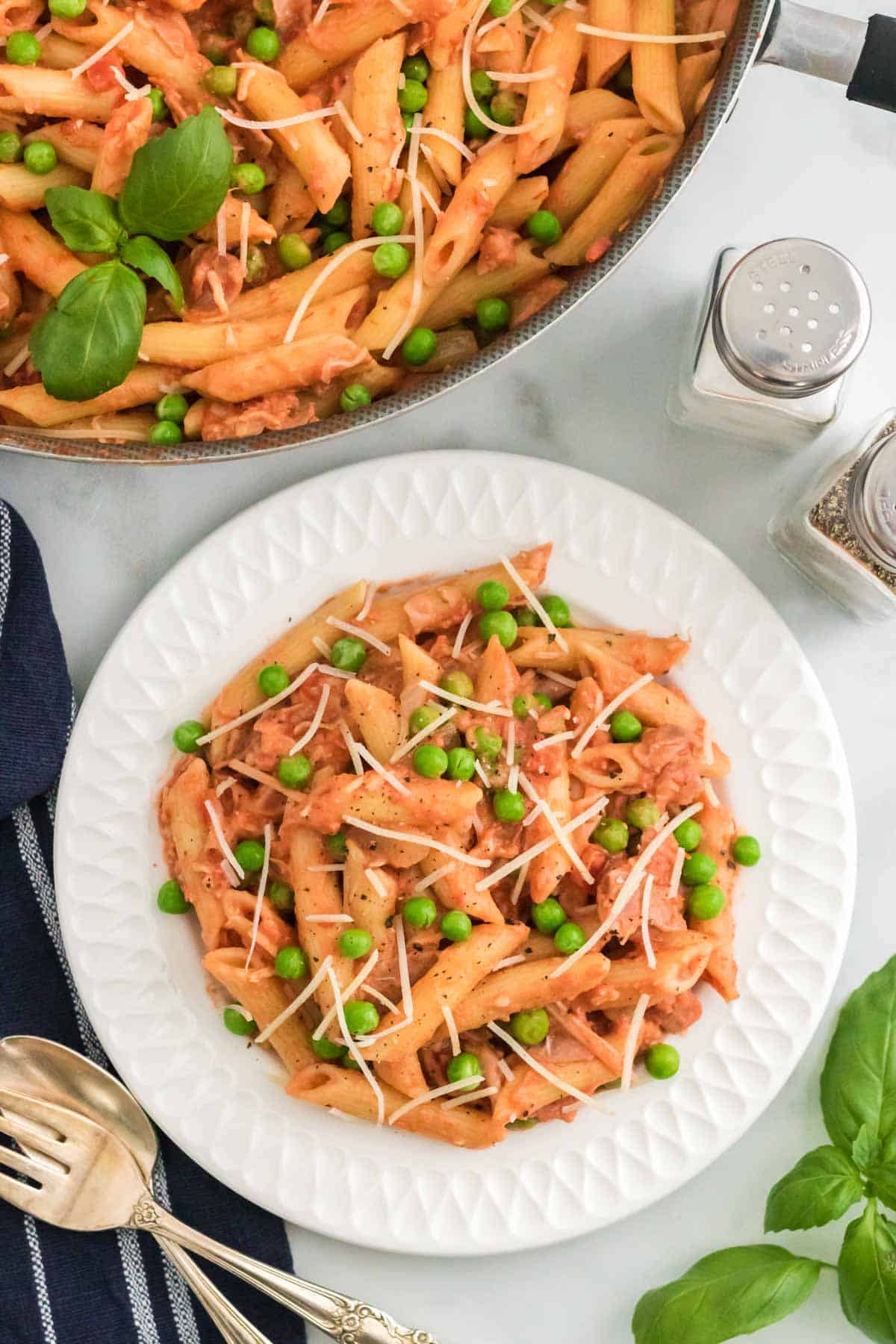 Penne with tomato cream sauce on a plate topped with peas and prosciutto, next to a pan of pasta.