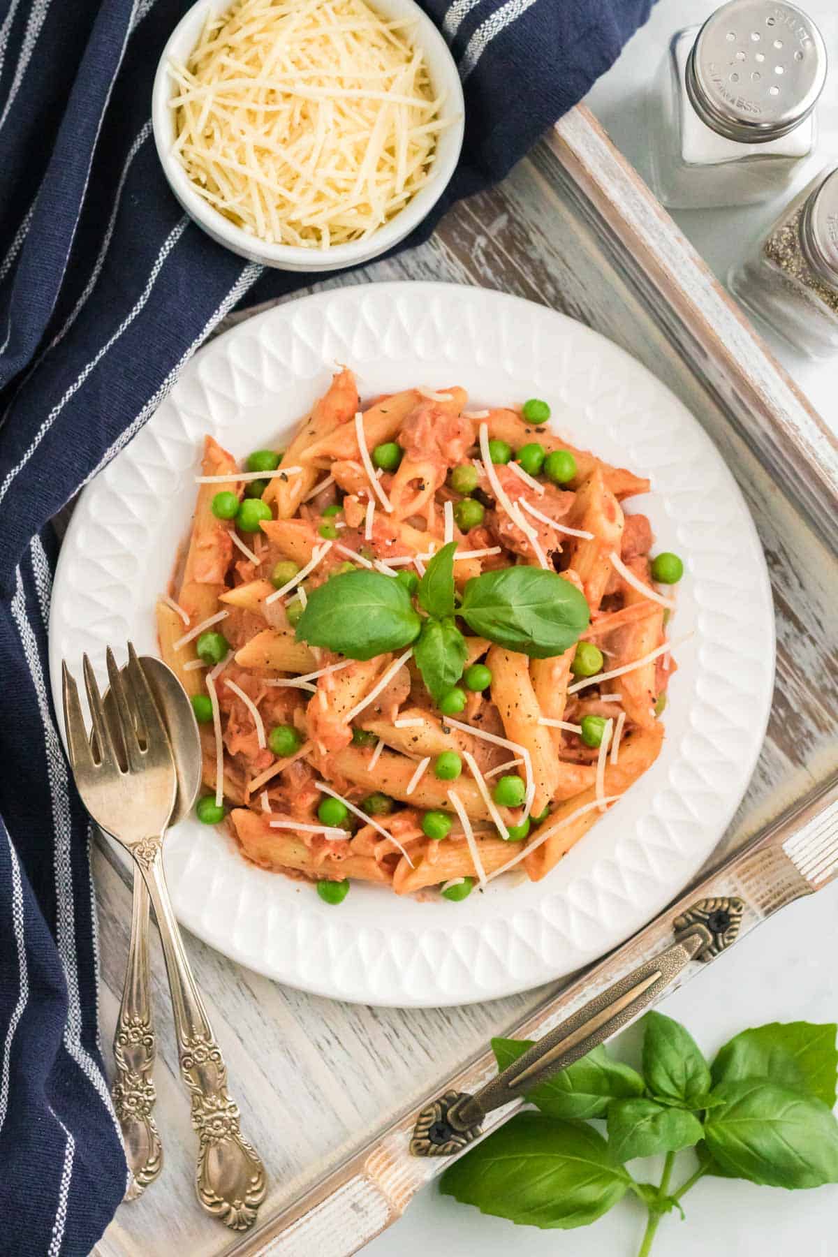 Penne with tomato cream sauce on a plate topped with peas and prosciutto, next to a fork and a bowl of grated parmesan.