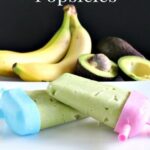 Two homemade avocado banana popsicles with pink and blue holders, with bananas and avocados in the background.