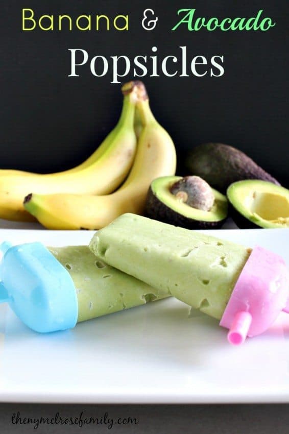 Two homemade avocado banana popsicles with pink and blue holders, with bananas and avocados in the background.