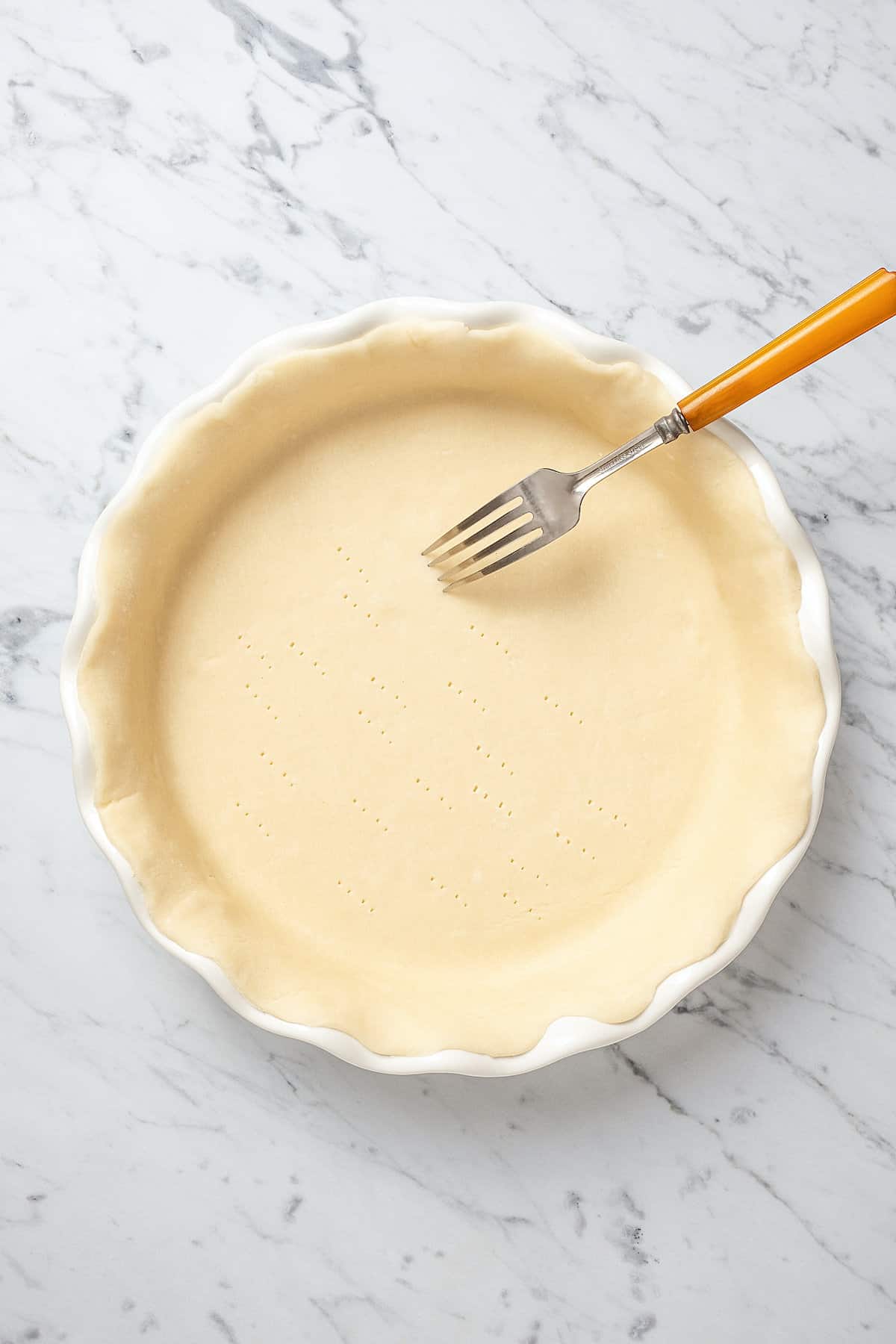 A fork pricks the bottom of a flaky pie crust in a pie plate.
