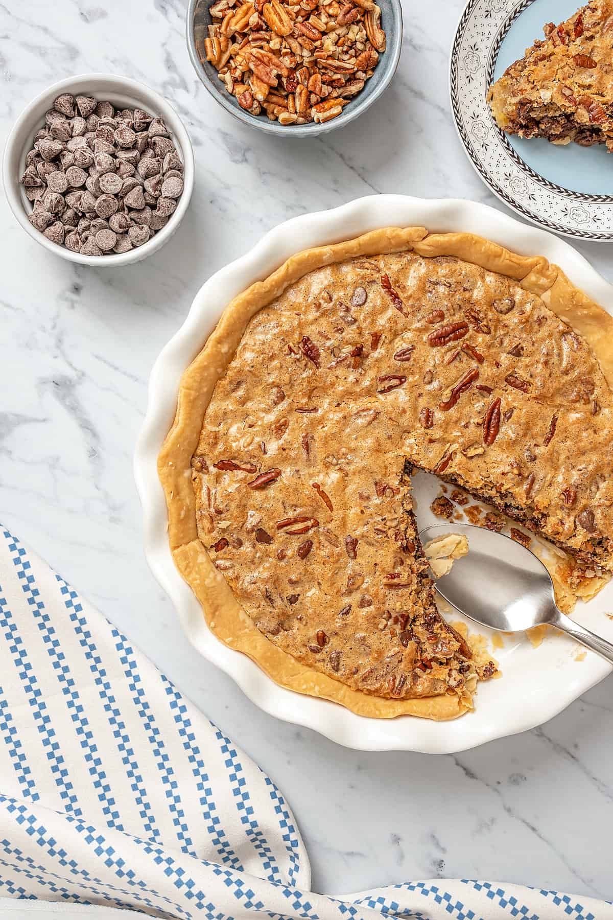 Gluten-free chocolate chip cookie pie with a slice missing, next to small bowls of chocolate chips and pecans.